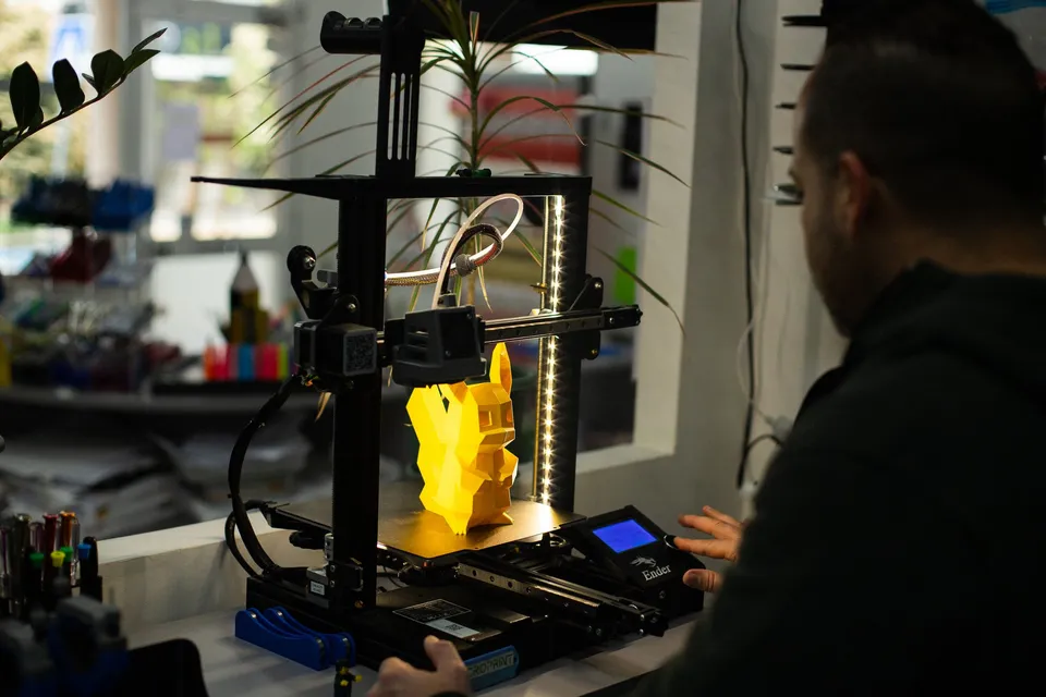 Impressora 3D a imprimir uma peça amarela, com um técnico ao lado